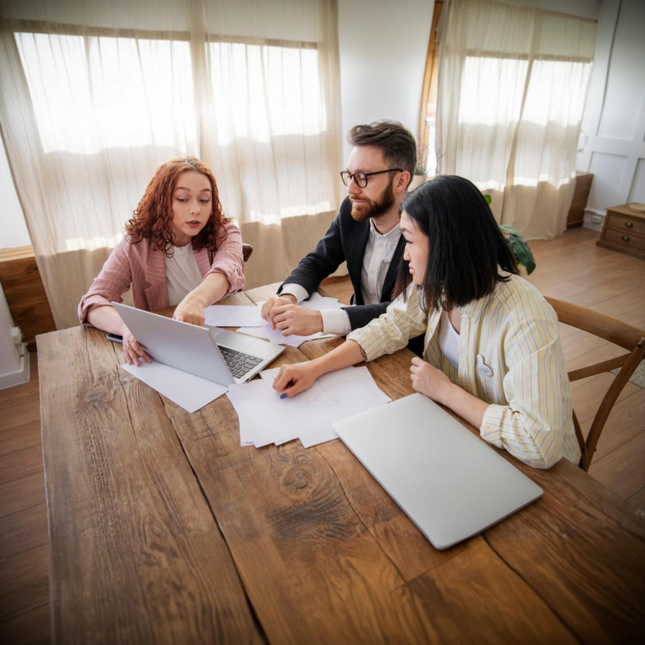 Three people sitting at a wooden table in a bright room, discussing documents and looking at a laptop together.