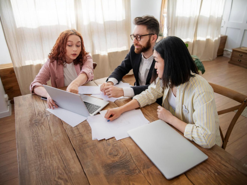 Three people sitting at a wooden table in a bright room, discussing documents and looking at a laptop together.